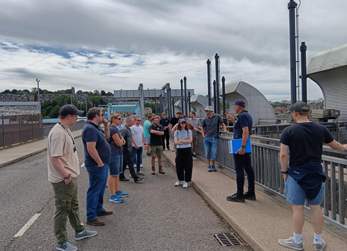 Photo of a group of people stood on a narrow road, listening to someone talking.