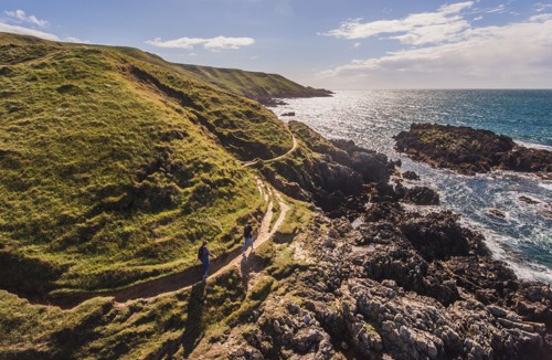 A Pair Of Walkers On The Wales Coastal Path At Porthor, Llŷn Peninsula Where Rugged Green Hills Meet The Sea