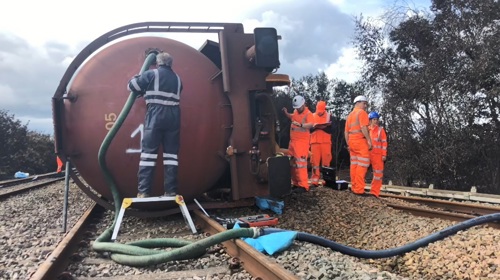 Train laying on it side after a derailment. Workers are pumping out contents of the tanker