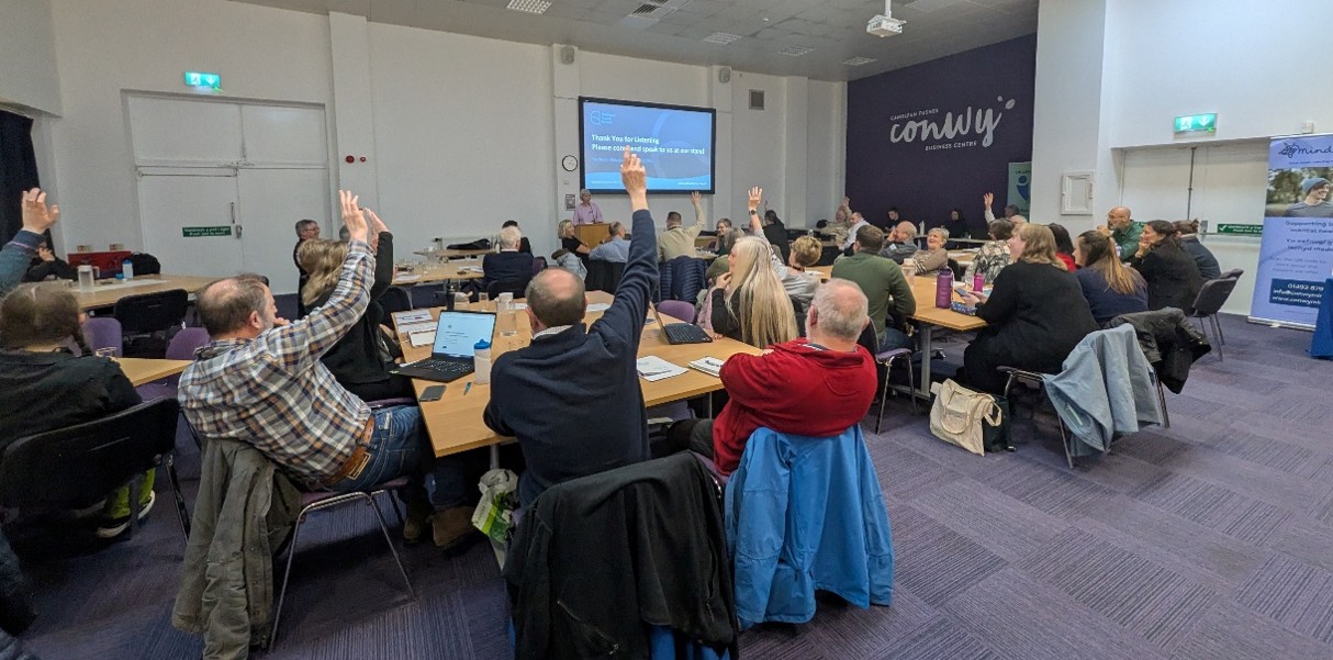 Photo of a room full of seated people, with a presentation on a large screen. Several people have their hands raised.