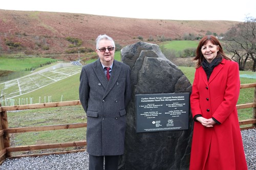Photo of the First Minister and Minster for Environment standing next to an engraved slate plaque on a large piece of stone from a local quarry, with the flood embankment and Dulais valley behind them.