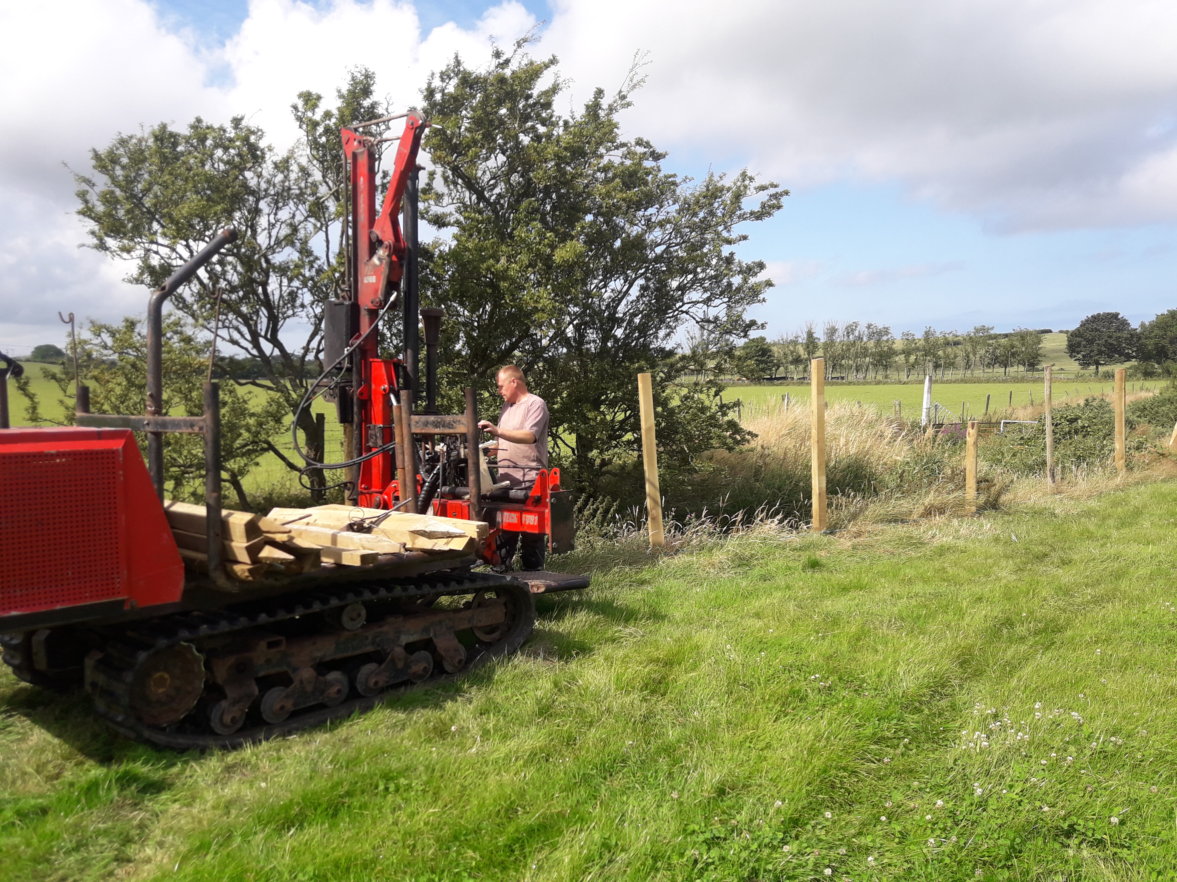 Photo of a man driving a large fence post machine next to Afon Wygyr with newly installed posts behind him