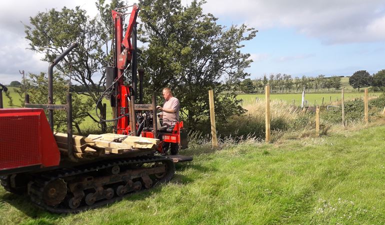 Photo of a man driving a large fence post machine next to Afon Wygyr with newly installed posts behind him