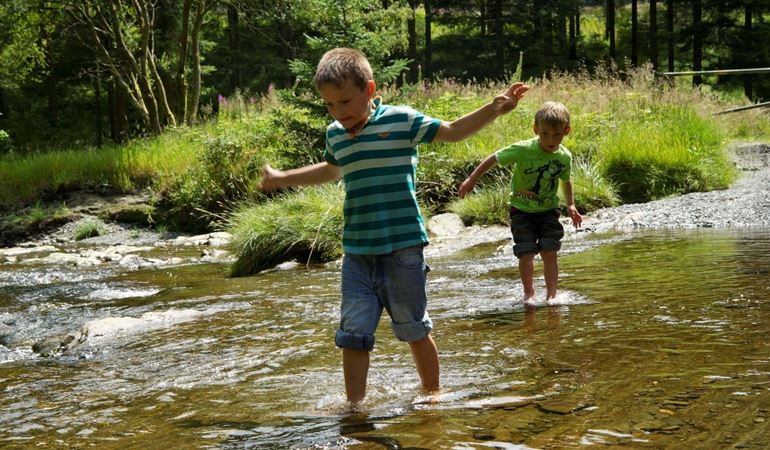 Two boys cross a shallow river barefoot.