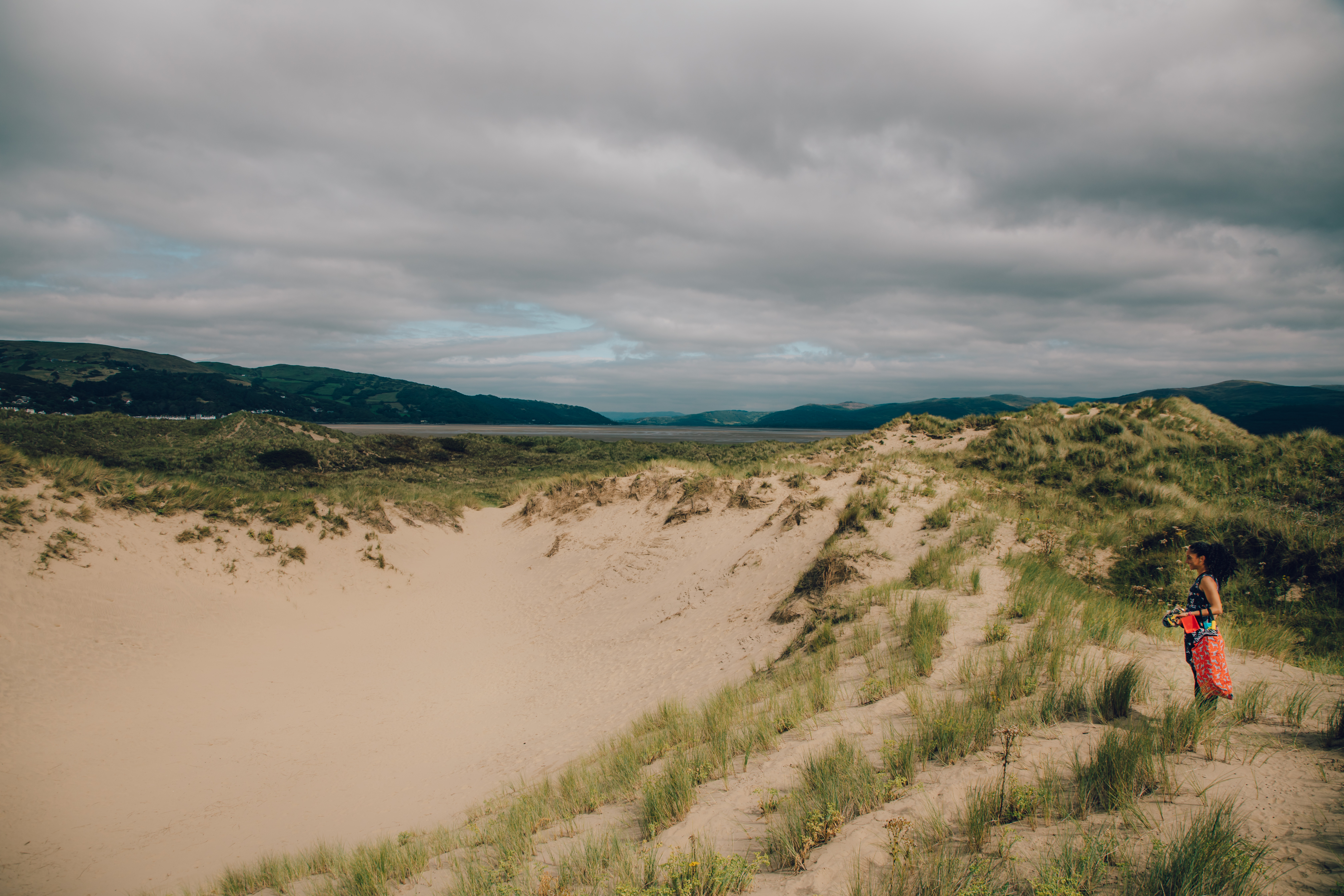 A woman standing next to a sand dune at Ynyslas, with the Dyfi Estuary in the background