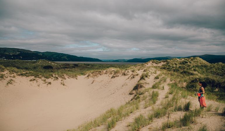 A woman standing next to a sand dune at Ynyslas, with the Dyfi Estuary in the background