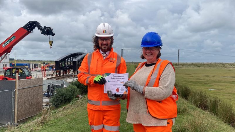Two people holding a signed agreement wearing safety clothing with construction machinery in the background Two people holding a signed agreement wearing safety clothing with construction machinery in the background
