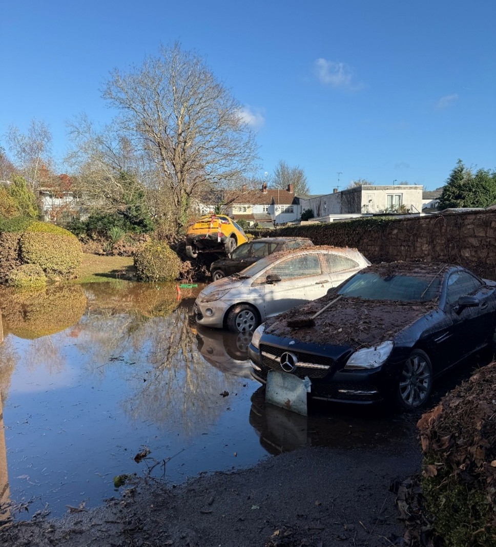 Photo of cars covered in thick mud and leaves after a severe flood, with a car in the background having tipped over